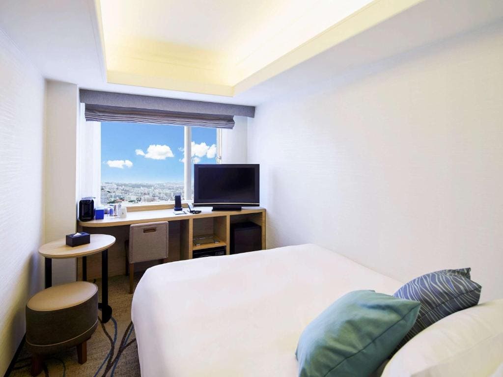 A modern hotel room with a large bed, two pillows, a small round table, a stool, a desk with a TV, a coffee maker, and a window showing a bright city view under a blue sky with clouds.