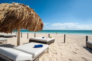 Lounge chairs with white cushions under a thatched umbrella on a sandy beach, overlooking turquoise ocean water and a clear blue sky. A person walks near the shoreline in the background.