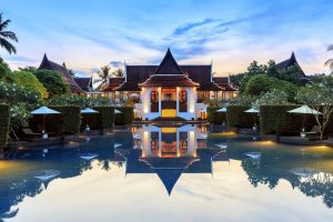 A luxurious building with traditional Thai architecture is reflected in a large, serene swimming pool, surrounded by manicured greenery and white umbrellas under a pastel blue evening sky.