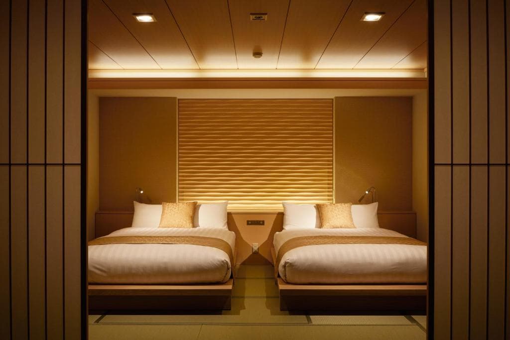 A modern hotel room with two double beds, white linens, beige bed runners, and matching pillows. The room features warm lighting, a wooden ceiling, and a textured wall behind the beds, creating a cozy, minimalist atmosphere.