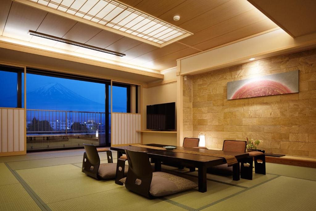 A traditional Japanese room with tatami mats, a low wooden table, floor chairs, and a large window offering a scenic view of Mount Fuji at dusk. Warm lighting and minimalist decor create a serene atmosphere.