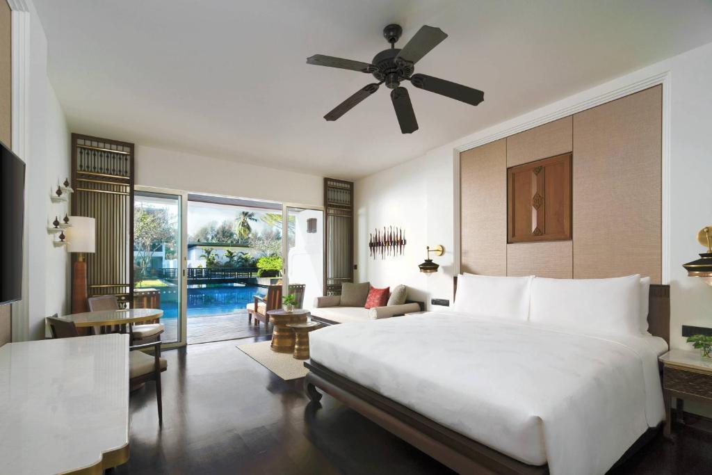 Modern hotel room with a large bed, sofa, desk, TV, and ceiling fan. Floor-to-ceiling windows open to a patio with a pool and green garden view. The room is bright and decorated in neutral tones.