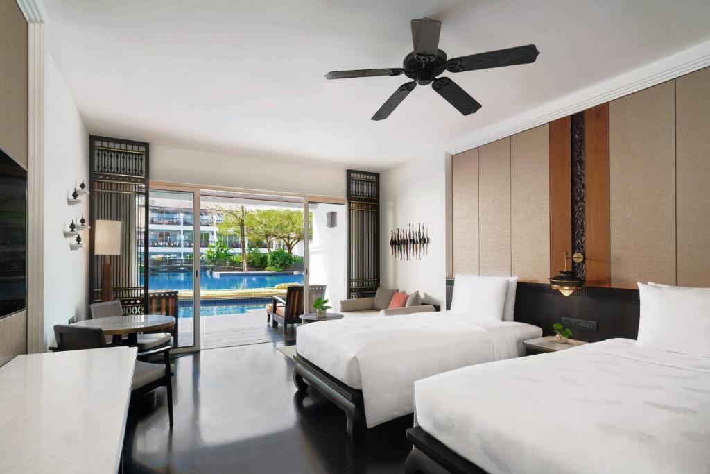Modern hotel room with two beds, a ceiling fan, a desk, a TV, and a seating area. Large sliding doors open to a poolside patio with a view of the pool and greenery outside. The decor is stylish and minimal.
