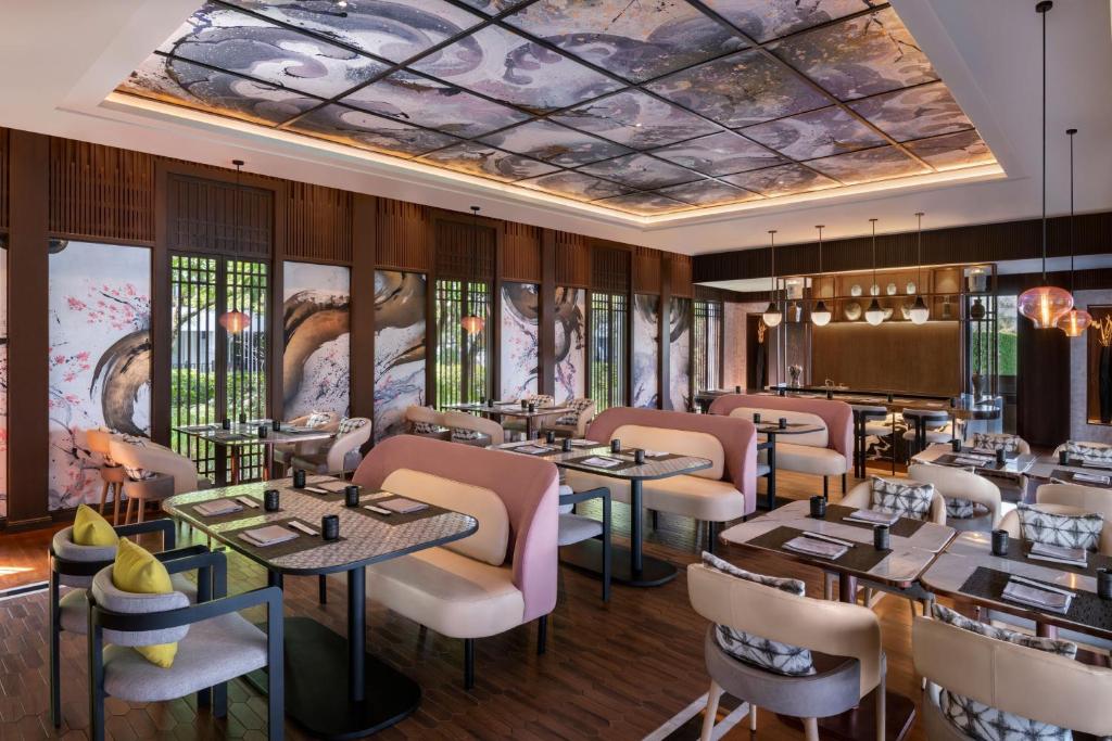 A modern restaurant with pink, beige, and gray seating, wooden flooring, large artistic wall panels, and a decorative ceiling. Tables are neatly set and soft lighting creates a cozy, elegant atmosphere.