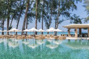 An inviting outdoor pool with lounge chairs, umbrellas, and rattan seating overlooks a sandy beach and turquoise ocean, with tall trees providing shade and a poolside bar nearby.