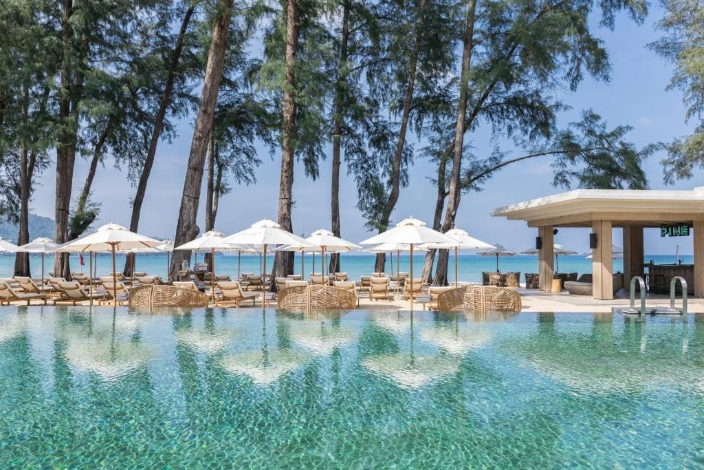 An inviting outdoor pool with lounge chairs, umbrellas, and rattan seating overlooks a sandy beach and turquoise ocean, with tall trees providing shade and a poolside bar nearby.