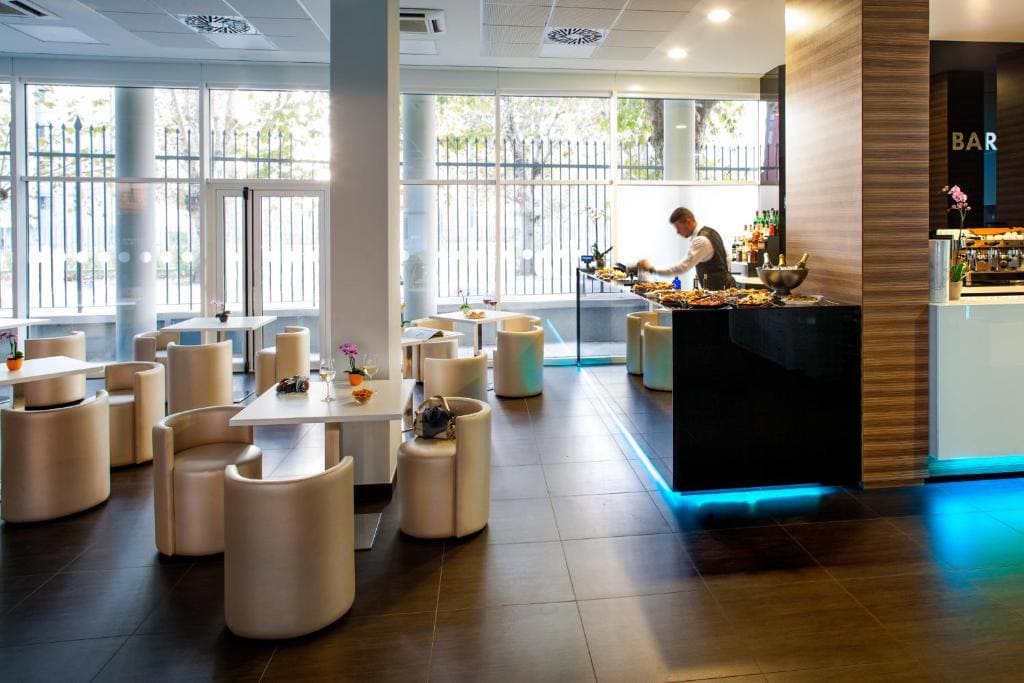 A modern, bright café with round white tables and chairs, large windows letting in natural light, and a bar area where a person arranges food on a counter with blue lighting underneath.