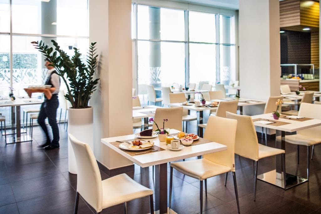 Bright, modern restaurant interior with white chairs and tables set for breakfast. A server carries a tray in the background. Large windows let in natural light; a tall potted plant stands near the center.