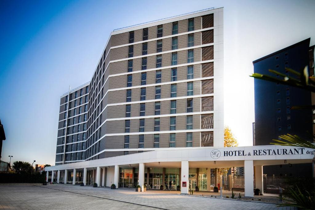 Modern multi-story hotel building with a geometric design, large windows, and a covered entrance. The sign reads Hotel & Restaurant. The sky is clear and trees are visible nearby.