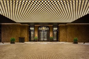 Luxurious hotel entrance with a grand, glass double door, flanked by two potted plants. The ceiling features a geometric grid of warm, glowing lights, and the floor is covered in elegant tiles.