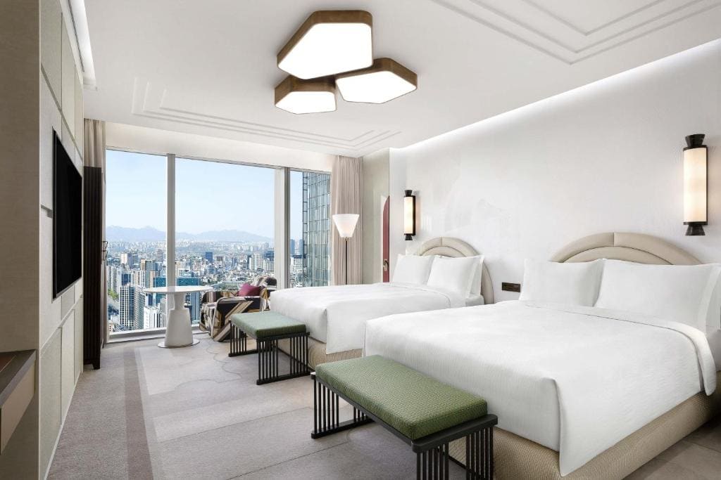 Modern hotel room with two large beds, green benches at the foot, a TV, floor-to-ceiling window showing a cityscape and mountains, and a person sitting near the window. The decor is minimalist and bright.