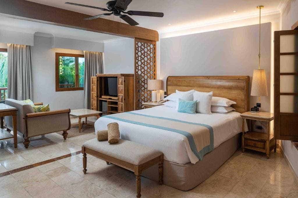 Spacious hotel room with a large bed, wooden furniture, a ceiling fan, soft lighting, lounge area with sofas, and windows overlooking greenery. Rolled towels are placed on a bench at the foot of the bed.