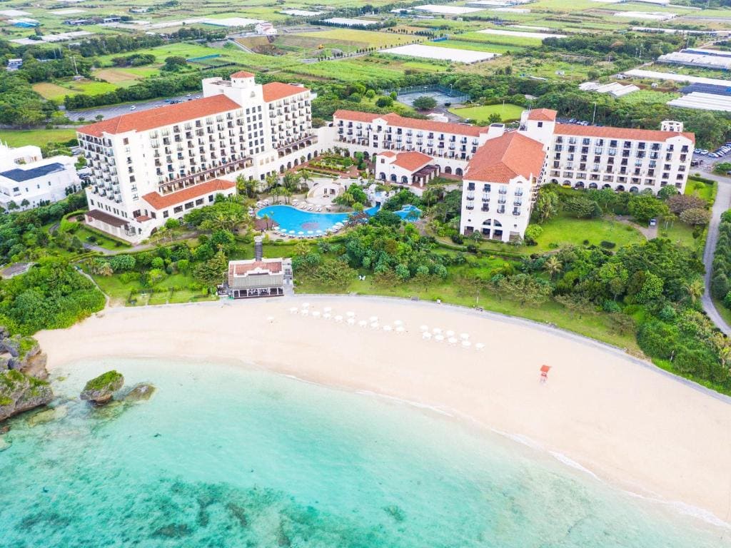 Aerial view of a large seaside resort with red-roofed buildings, a central pool, lush greenery, and a sandy beach with rows of umbrellas next to turquoise water. Fields and roads are visible in the background.