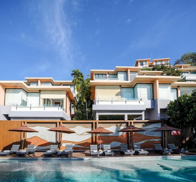 Modern resort buildings with large windows overlook a swimming pool lined with sun loungers and brown umbrellas, set against a clear blue sky and surrounded by greenery.