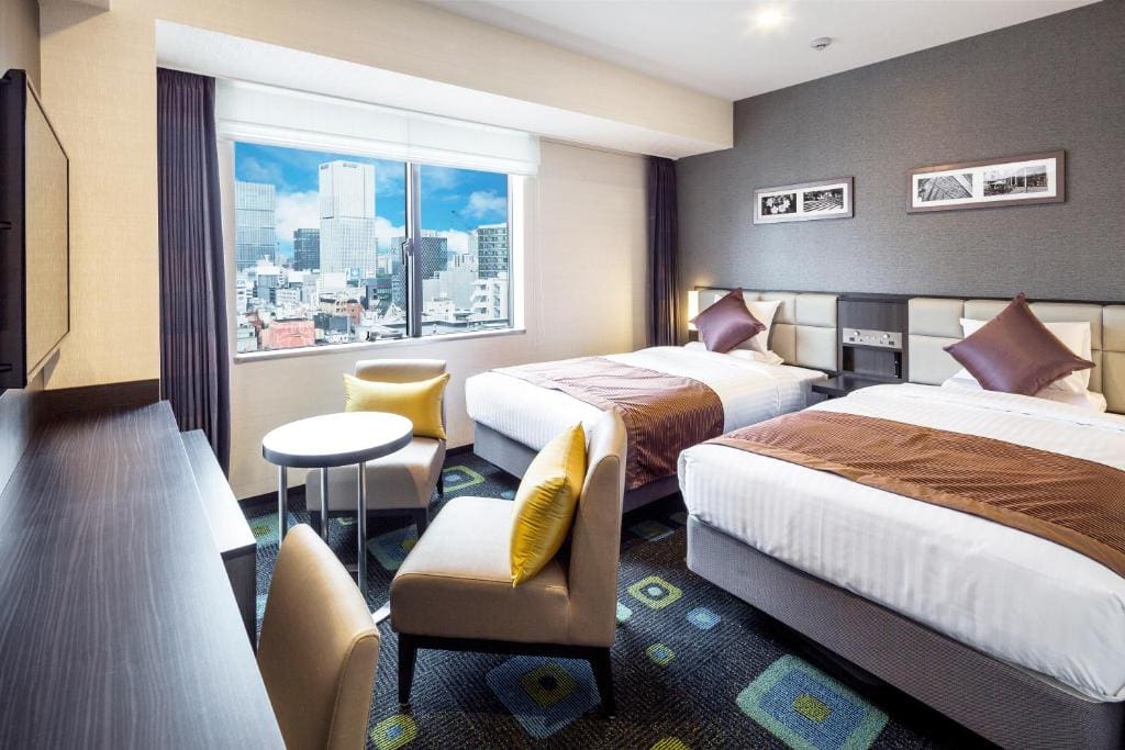 Modern hotel room with two double beds, a large window showing a cityscape, a small round table with two armchairs, a long desk, and wall art above the beds. The décor is neutral with brown and yellow accents.