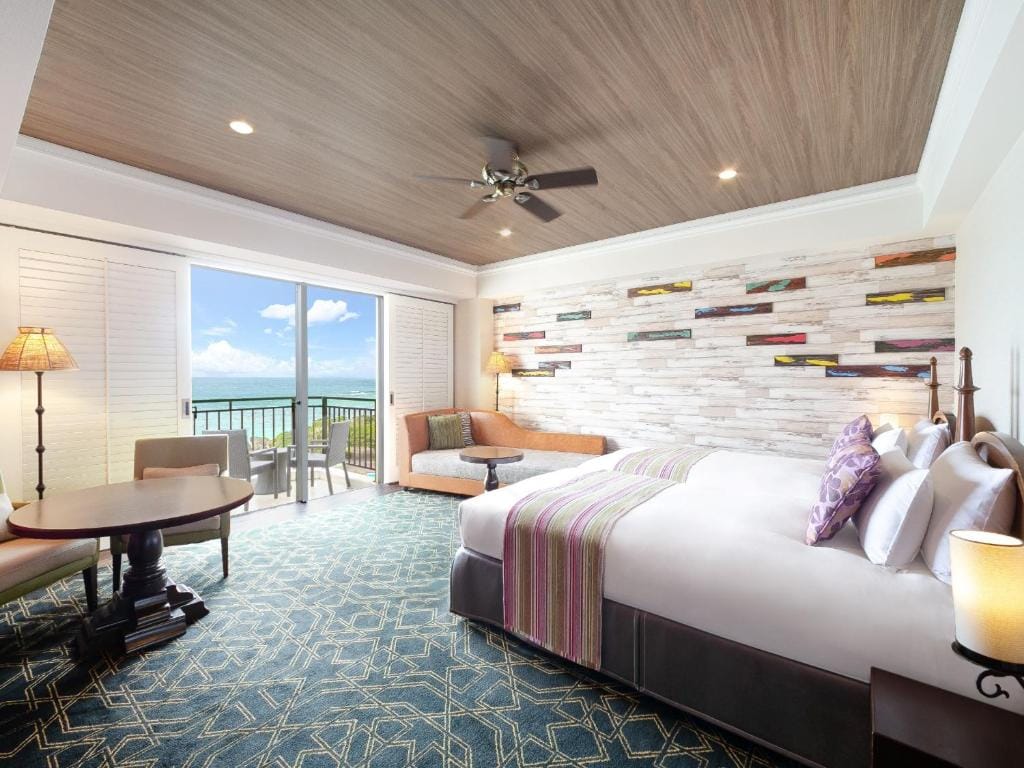 A spacious hotel room with a large bed, a sofa, a table with chairs, and a balcony overlooking the ocean. The room features modern decor, a wooden ceiling, and a wall with colorful wood accents.
