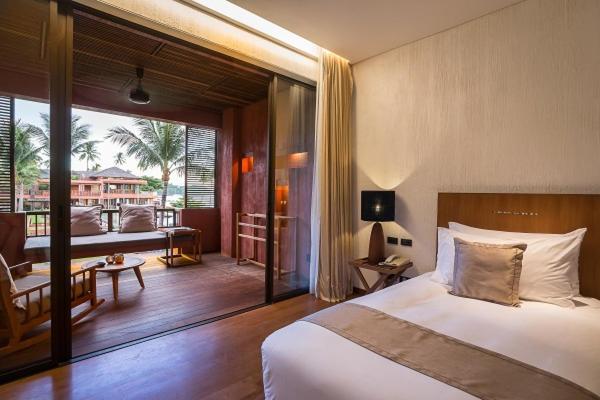 A modern hotel room with a neatly made bed, bedside table, lamp, and sliding glass doors opening to a balcony with seating. Palm trees and another building are visible outside. Warm lighting creates a cozy atmosphere.