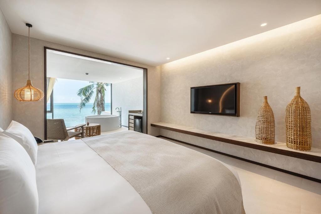 Modern hotel room with a large bed, neutral decor, wall-mounted TV, wicker accents, and a balcony featuring a freestanding bathtub, chairs, and an ocean view with a palm tree.