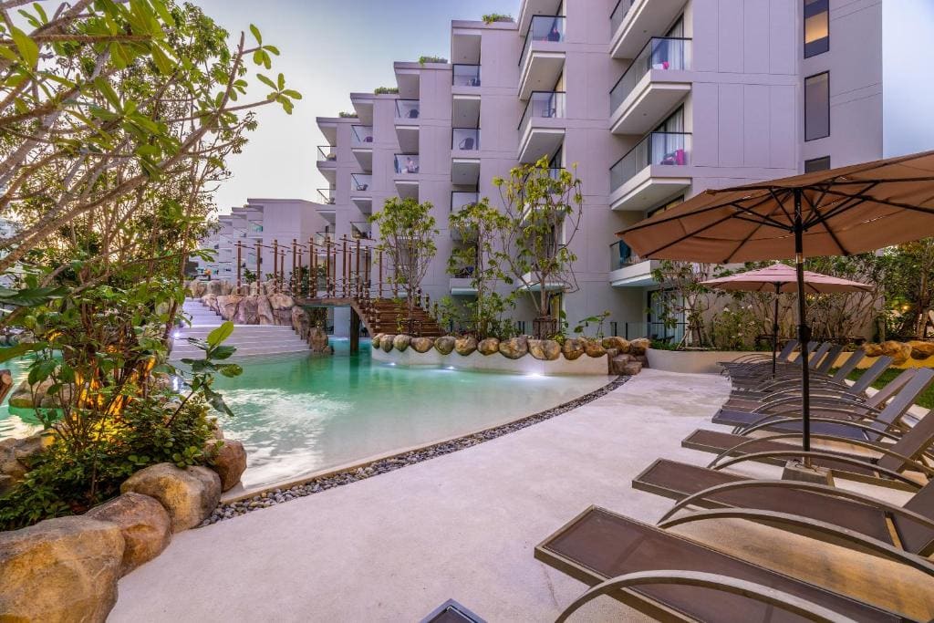 A modern hotel pool area with lounge chairs, large umbrella, lush greenery, and a bridge over a clear turquoise pool beside a contemporary multi-story building.