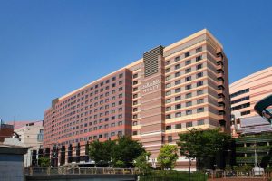 The image shows a large, multi-story Grand Hyatt hotel building with reddish-brown and beige horizontal stripes, set against a clear blue sky and surrounded by green trees.