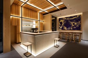 Modern minimalist cafe with light wood accents, a textured L-shaped counter, two stools, a coffee machine, a large abstract blue and white artwork, and tables with chairs along the back wall. Warm ambient lighting throughout.