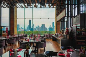 A modern restaurant with tables set for dining, large floor-to-ceiling windows offering a scenic view of a city skyline with tall buildings and trees outside. Natural light fills the space, creating a vibrant atmosphere.