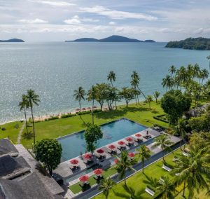 Aerial view of a tropical beachfront resort with a large swimming pool, palm trees, green lawn, and red umbrellas; calm blue sea and distant islands under a sunny sky.