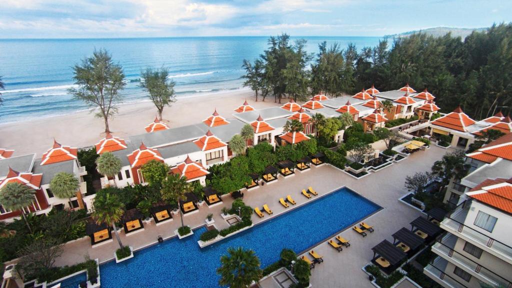 Aerial view of a beachfront resort with orange-roofed villas, a large rectangular swimming pool, yellow lounge chairs, lush greenery, and the ocean with gentle waves and sandy beach in the background.