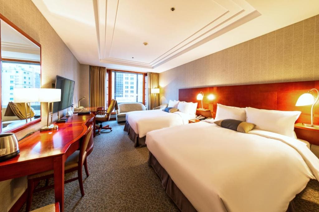 Bright hotel room with two large beds, a desk with a chair, a TV, lamps, a full-length mirror, and a window with city views. The decor is modern and welcoming.