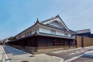 Traditional Japanese building with white walls, dark wooden accents, and a tiled roof, situated on a quiet, empty street with a wooden fence and clear blue sky overhead.