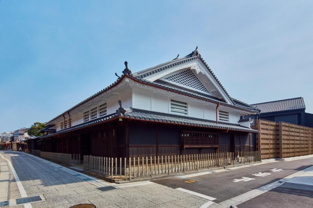 Traditional Japanese building with white walls, dark wooden accents, and a tiled roof, situated on a quiet, empty street with a wooden fence and clear blue sky overhead.