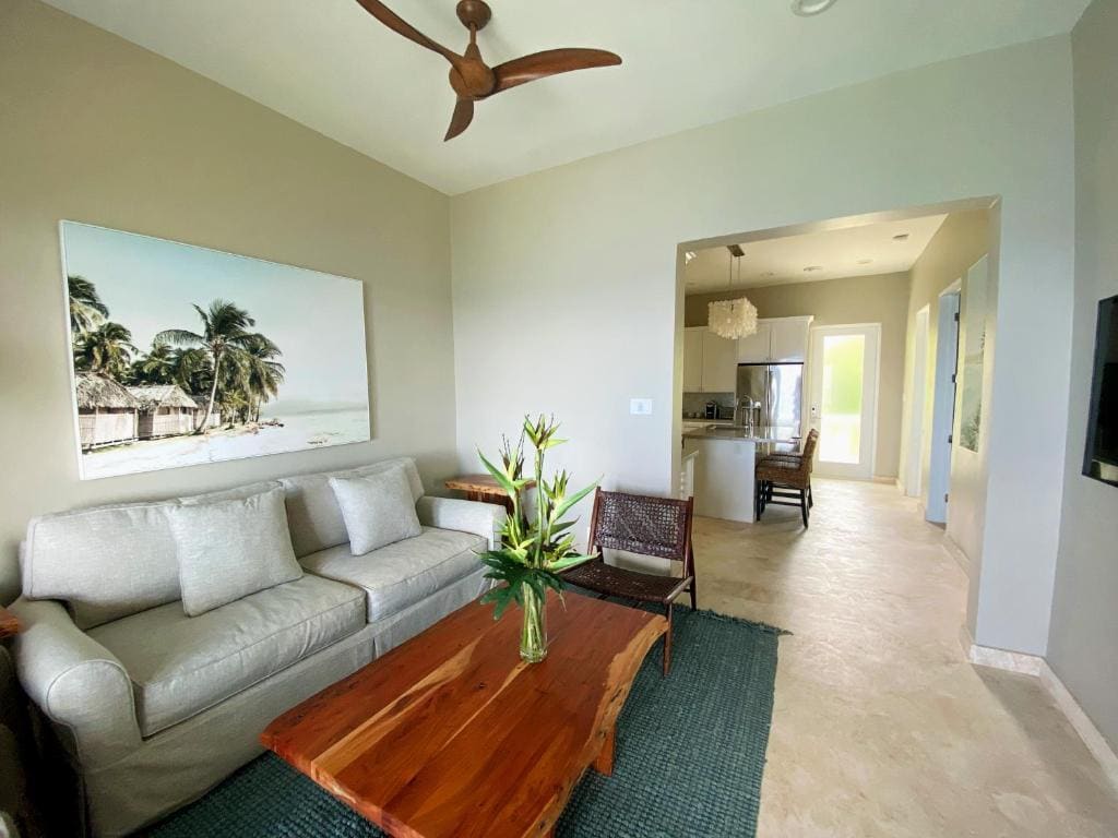 A modern living room with a light gray sofa, a wooden coffee table with a flower arrangement, a tropical beach photo on the wall, and an open view into a kitchen with neutral tones.