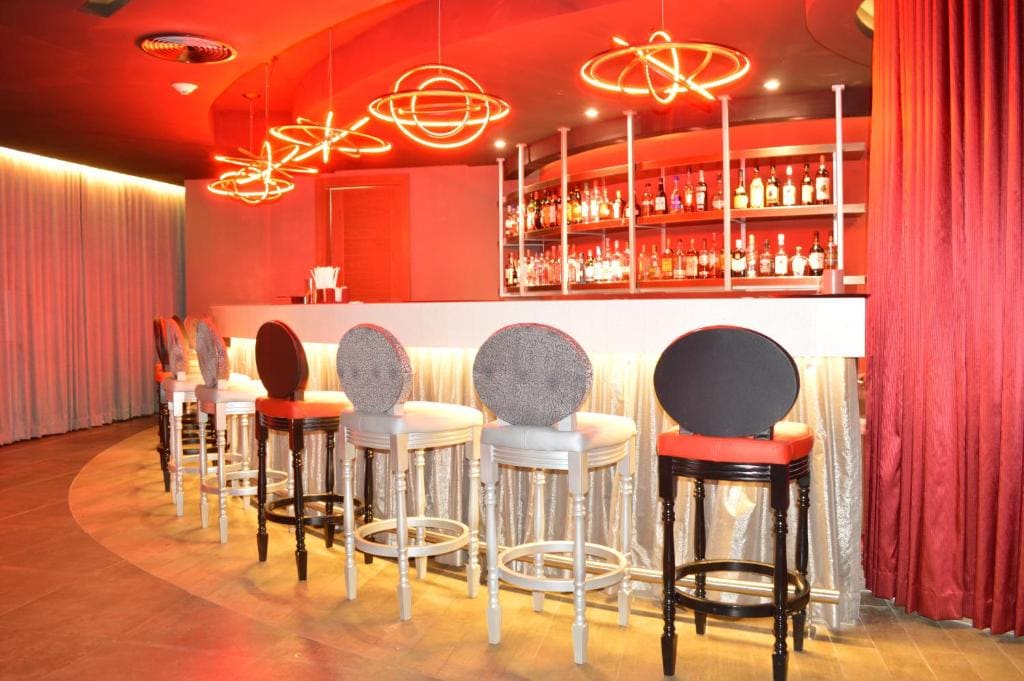 A modern bar with a white counter, high-backed stools, red and gray decor, and shelves stocked with bottles. Neon red circular lights hang above, creating a vibrant, stylish atmosphere.