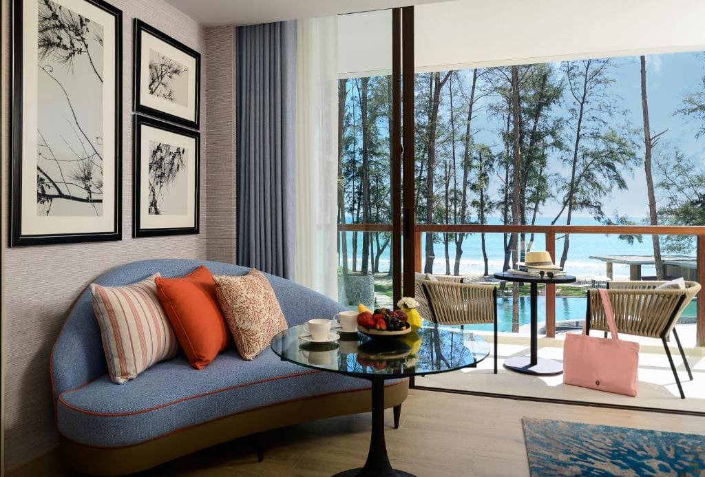 A modern hotel room with a curved blue sofa, colorful pillows, a round glass table with fruit and drinks, and a balcony overlooking a pool, trees, and the ocean under a clear sky.