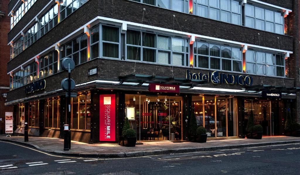 A modern corner building with large windows and warm interior lighting houses the Hotel Indigo. The entrance displays signs for Square Mile and the hotel, with potted plants flanking the doors.