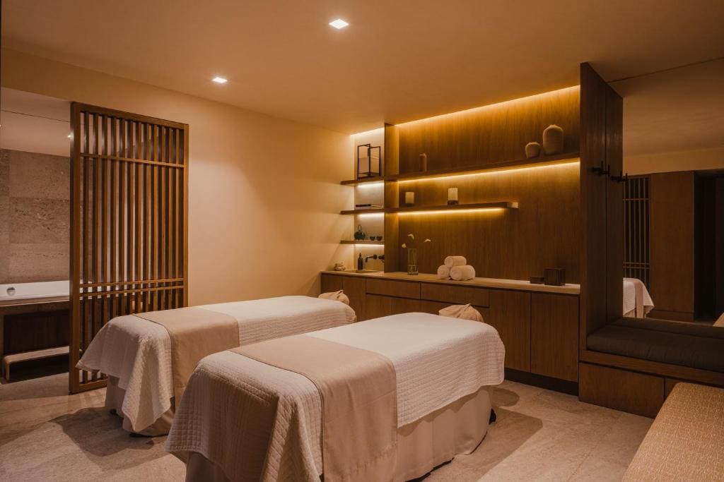 A tranquil spa room with two massage tables covered in white linens, soft lighting, wooden shelves with rolled towels and decor, and a peaceful, minimalist atmosphere.