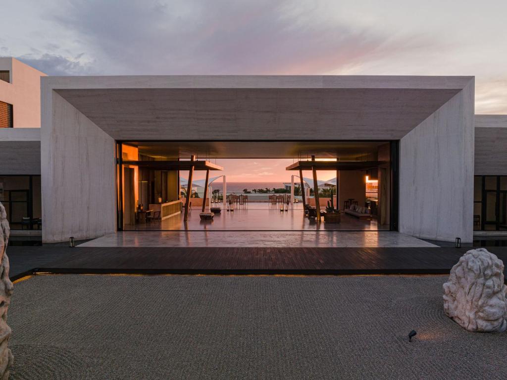 A modern, open-air building with a wide entrance frames a view of a pool and palm trees at sunset, reflecting soft pink and orange hues in the sky. The foreground features a gravel garden with stone accents.