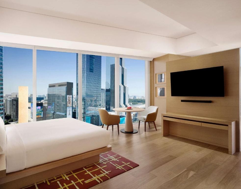 A modern hotel room with a large bed, a wall-mounted TV, a small dining table with two chairs by floor-to-ceiling windows, and a city skyline view outside. The room has light wood floors and minimalist decor.