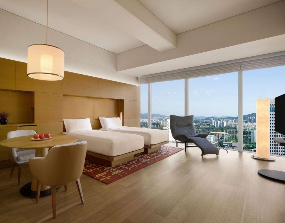 A modern hotel room with two beds, a round table with chairs, a lounge chair, and floor-to-ceiling windows offering a cityscape view. The room has wooden floors and minimalist décor.