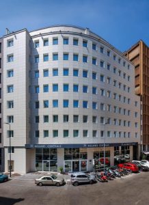 Modern multi-story hotel named “Milano Centrale” with a curved white facade, large windows, and ground-floor glass entrance. Several cars and motorcycles are parked along the street in front of the building under a clear sky.