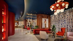A modern hotel lobby with mid-century furniture, bold red and orange chairs, gold accents, abstract art, wooden paneling, decorative shelving, potted plants, and geometric-patterned curtains.