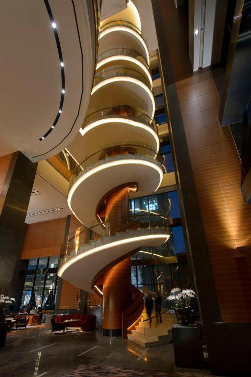 A modern hotel lobby featuring a dramatic, spiral staircase with glowing lights, surrounded by tall glass windows, sleek dark floors, and wooden panel accents. Several people stand near the staircase base.