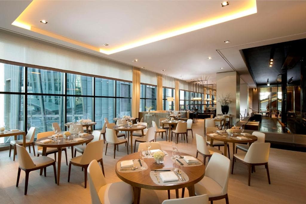A modern, upscale restaurant with round wooden tables set for dining, cream-colored chairs, large floor-to-ceiling windows, and soft ambient lighting. The space is clean, bright, and stylish with minimalist decor.