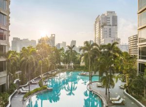 A serene outdoor pool surrounded by palm trees and lounge chairs, set amidst tall modern buildings with the sun rising or setting in the background.