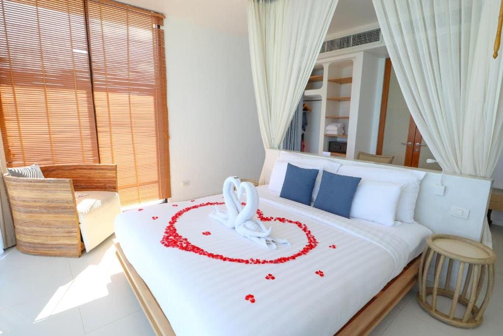A neatly made bed with white linens, blue accent pillows, and rose petals arranged in a heart shape. Two towel swans form another heart in the center. The room has wooden furniture, sheer curtains, and natural light.