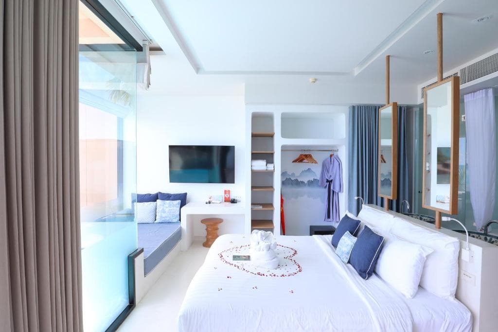 A bright, modern hotel room with a large bed decorated with rose petals, a sofa by the window, a mounted TV, open wardrobe with a robe, and glass doors leading to a pool area outside.
