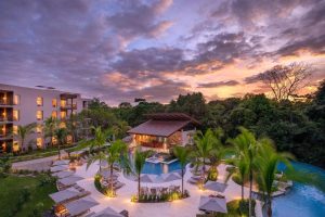 Lush tropical resort with a glowing pool and palm trees at sunset, surrounded by greenery. Lounge chairs and umbrellas are arranged around the pool, and a modern building stands to the left under a dramatic, colorful sky.