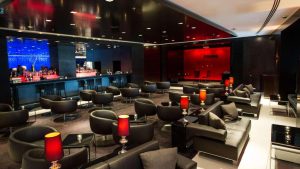 A modern lounge bar with black chairs and sofas, red lamps on tables, and a sleek bar area in the background illuminated with blue and red lighting. The space has a stylish, contemporary design.