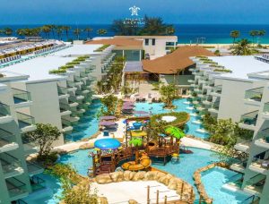 Aerial view of a resort with multiple white buildings, winding pools, water slides, play areas, palm trees, and a beach with blue ocean in the background. Phuket Emerald logo is visible at the top.