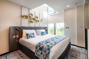 A modern hotel room with a large bed, geometric-patterned bedding, unique gold wall art above the headboard, and a window letting in natural light with a view of trees outside.